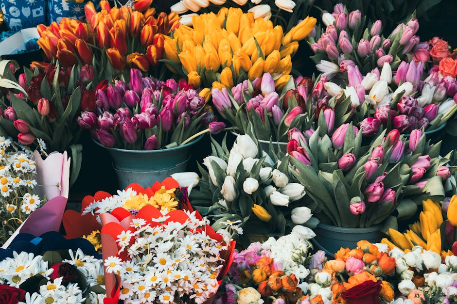 A vibrant display of fresh tulips in various shades, including pink, white, yellow, and orange, arranged in rustic metal buckets and along a table. The tulips have smooth, elongated petals and sturdy 