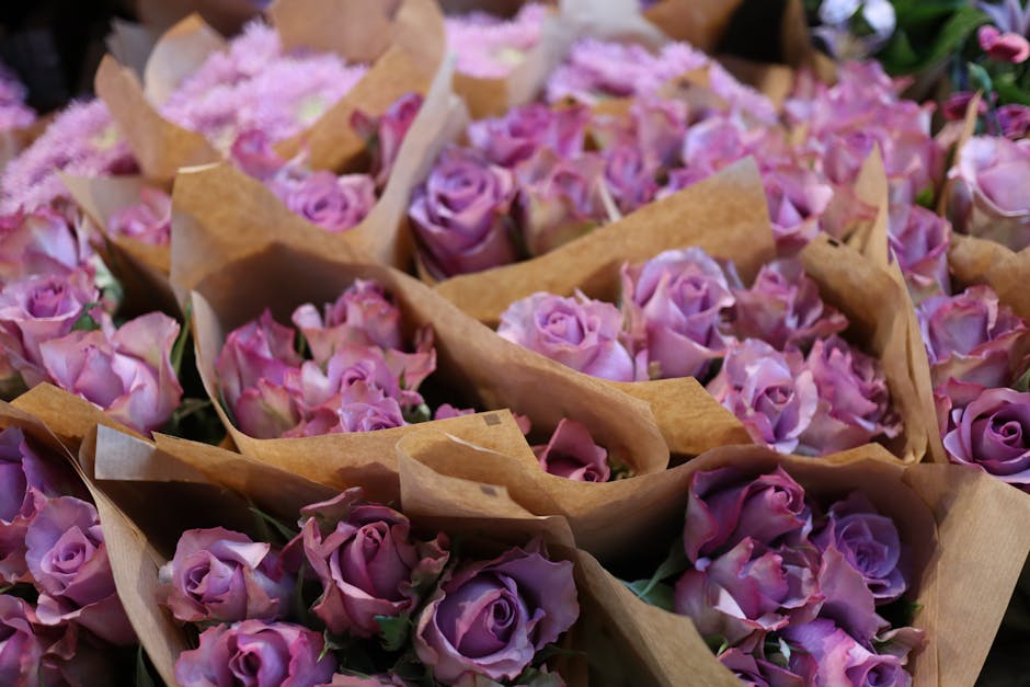 A collection of fresh pink and lavender roses arranged in small bouquets, each wrapped in light brown craft paper. The roses have tightly packed petals with a soft texture, and vary slightly in shades