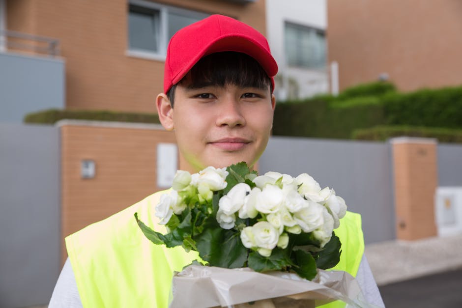 A young man wearing a red cap and a high-visibility yellow jacket holding a floral bouquet featuring white roses and green foliage. The bouquet is wrapped in white paper and is being delivered outdoor
